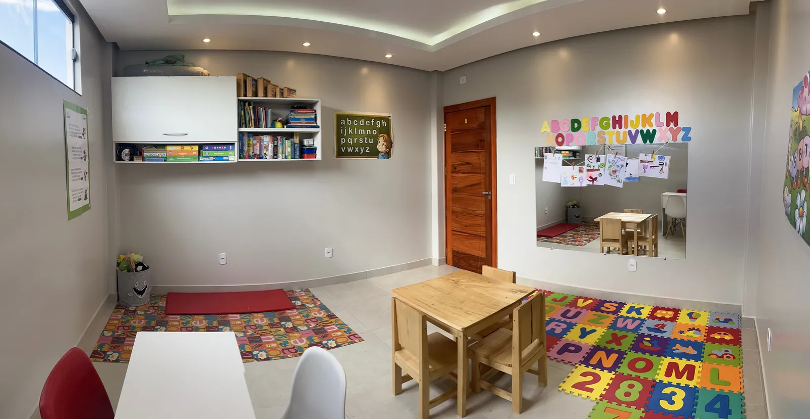 Sala de neuropsicopedagogia, com tapete de letras e números em EVA, mesinha de madeira e estante de jogos.