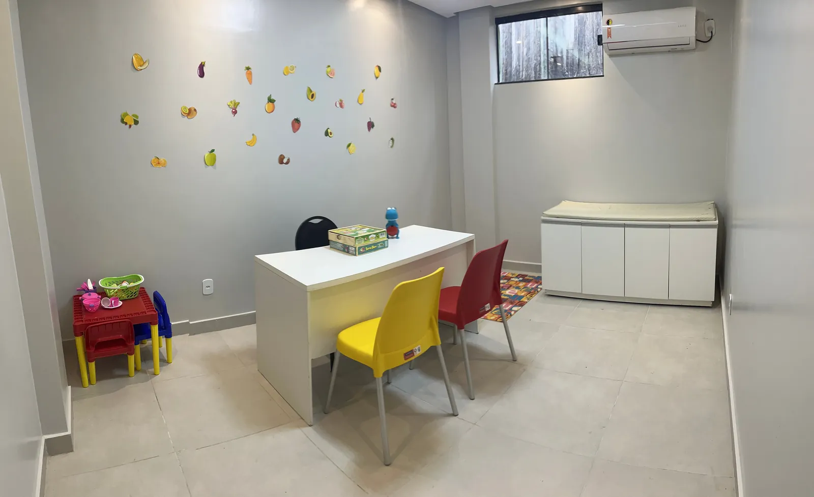 Consultório de nutrição, com mesa, cadeiras coloridas e uma mesinha infantil com comidinhas de brinquedo.
